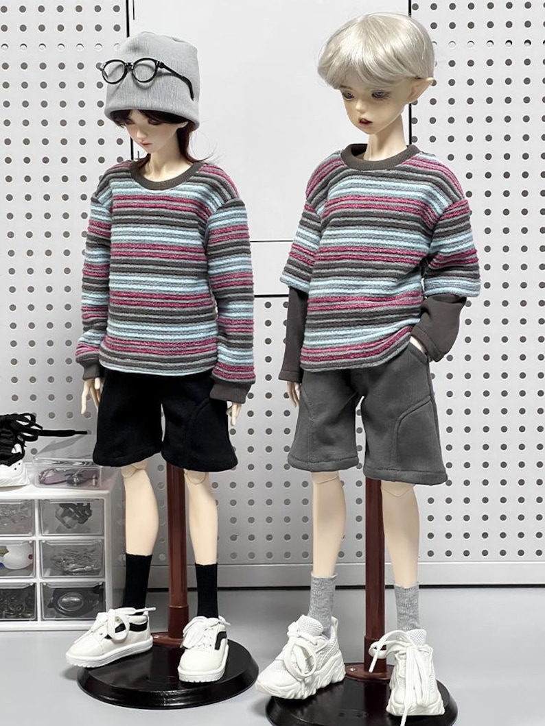 Pu&ograve; includere: Due bambole che indossano camicie a maniche lunghe a righe e pantaloncini. La bambola a sinistra indossa un berretto grigio e occhiali. Entrambe le bambole indossano scarpe da ginnastica bianche.