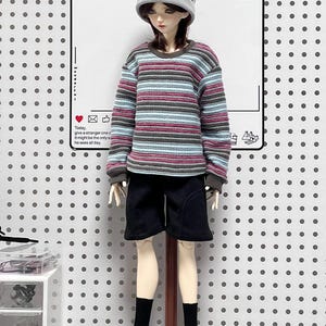 Pu&ograve; includere: Una bambola che indossa un berretto grigio, occhiali con montatura nera, una camicia a maniche lunghe a righe, pantaloncini neri, scarpe da ginnastica bianche e calzini neri. La bambola &egrave; in piedi su un supporto di legno.