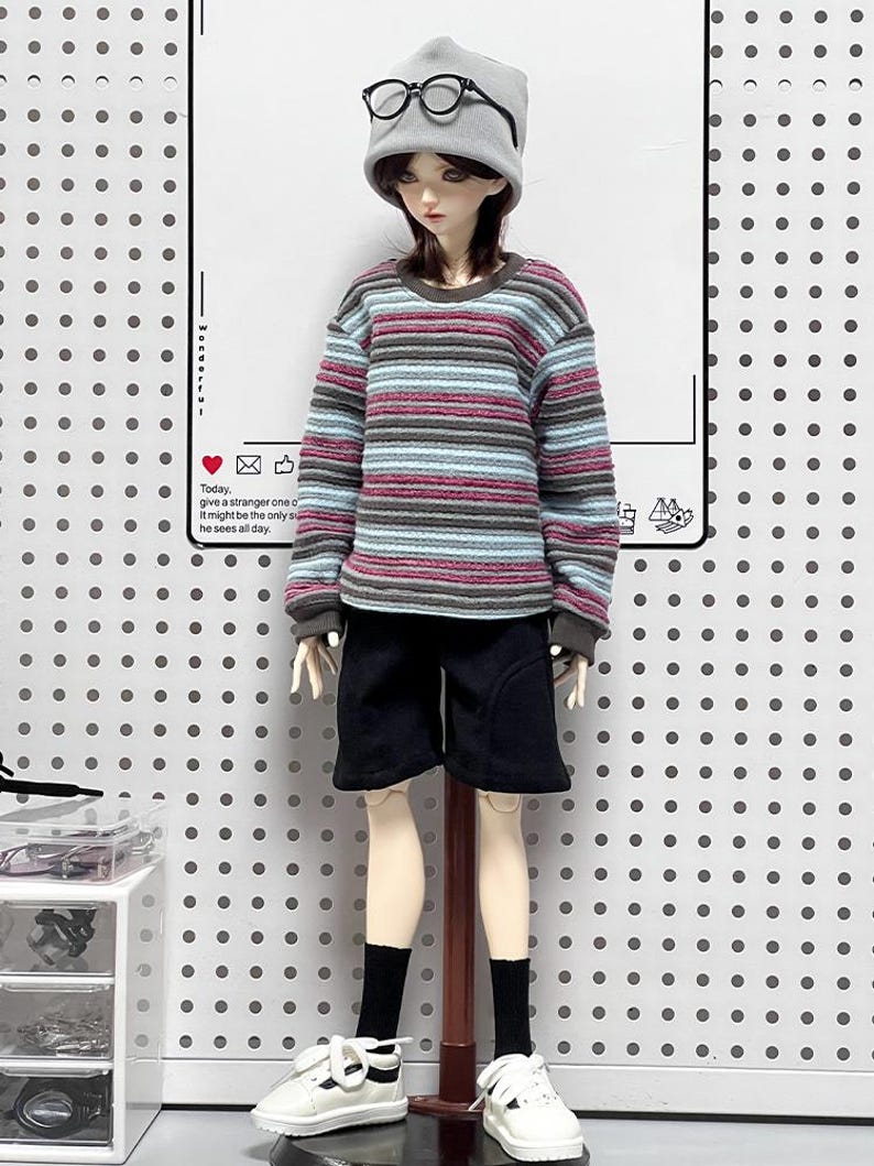 Pu&ograve; includere: Una bambola che indossa un berretto grigio, occhiali con montatura nera, una camicia a maniche lunghe a righe, pantaloncini neri, scarpe da ginnastica bianche e calzini neri. La bambola &egrave; in piedi su un supporto di legno.