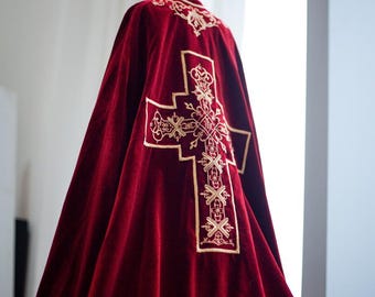 Op de afbeelding: Een dieprode fluwelen cape met gouden borduursel. De cape heeft een groot, sierlijk kruisontwerp op de rug en decoratieve patronen langs de randen. Het kledingstuk wordt waarschijnlijk gebruikt voor religieuze of ceremoniële doeleinden.