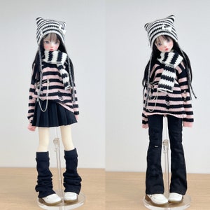 May include: Two dolls wearing black and white striped sweaters, black pants, and black and white striped hats. Both dolls are wearing black and white striped scarves. The doll on the left is wearing a black skirt and black knee-high socks. The doll on the right is wearing black flared pants. Both dolls are standing on white stands.