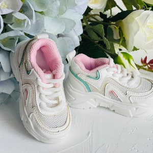 Puede incluir: Zapatillas blancas con detalles en rosa y azul. Las zapatillas tienen una parte superior de malla y una suela gruesa.
