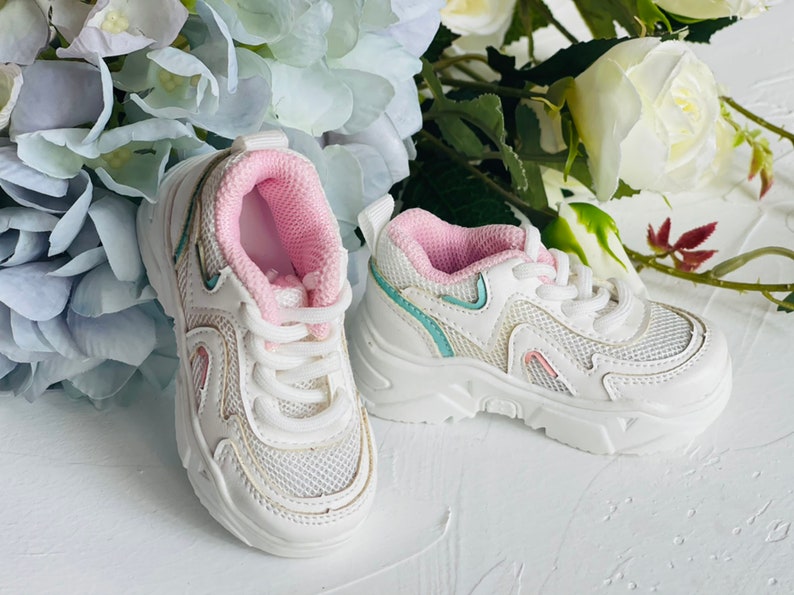 Puede incluir: Zapatillas blancas con detalles en rosa y azul. Las zapatillas tienen una parte superior de malla y una suela gruesa.