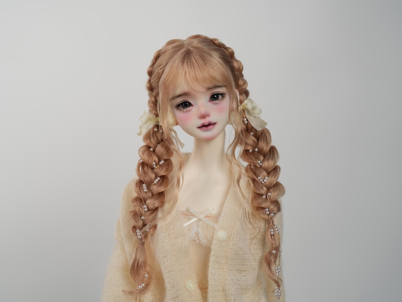 May include: A doll with a pale complexion, rosy cheeks, and full lips. The doll has long, light brown hair styled in two thick braids adorned with small pearls. She wears a cream-colored sweater with a bow and button details.