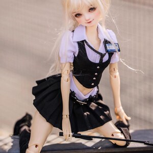 May include: A poseable doll with long, blonde hair, wearing a school uniform consisting of a white collared shirt, black vest, pleated skirt, and black shoes. The doll has a name tag and is holding a black rod.