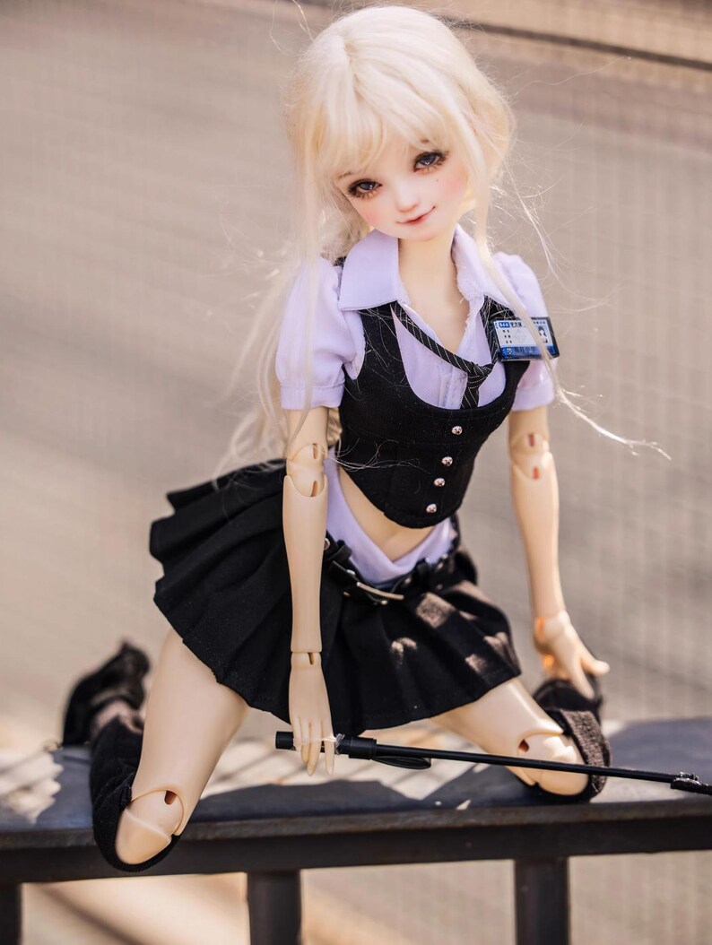 May include: A poseable doll with long, blonde hair, wearing a school uniform consisting of a white collared shirt, black vest, pleated skirt, and black shoes. The doll has a name tag and is holding a black rod.