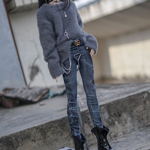 May include: A doll dressed in a grey fluffy sweater, dark wash jeans, and black boots with gold-coloured toes. The doll has black hair and wears a choker necklace. A small chain hangs from the belt. A small, fluffy, spotted bag is on the ground.