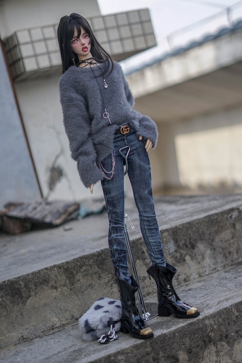 May include: A doll dressed in a grey fluffy sweater, dark wash jeans, and black boots with gold-coloured toes. The doll has black hair and wears a choker necklace. A small chain hangs from the belt. A small, fluffy, spotted bag is on the ground.