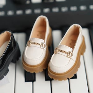 May include: A pair of white leather loafers with gold buckles. The loafers have a chunky brown sole and are sitting on a white and black surface.