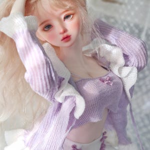 May include: A doll with long blonde hair, wearing a light purple knitted top and matching cardigan with white trim. The doll's skirt is white with purple ribbon detailing. The doll has a delicate face with rosy cheeks and lips.