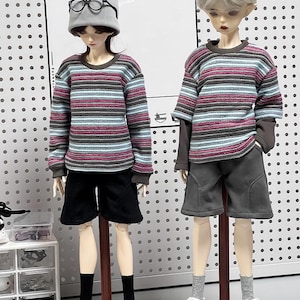 Pu&ograve; includere: Due bambole che indossano camicie a maniche lunghe a righe e pantaloncini. Una bambola indossa un berretto di lana grigio e occhiali. Entrambe le bambole indossano scarpe da ginnastica bianche.