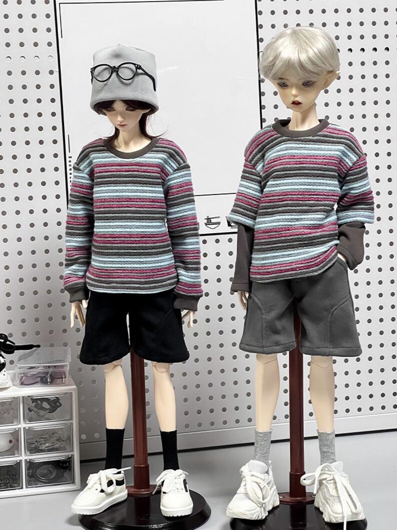 Pu&ograve; includere: Due bambole che indossano camicie a maniche lunghe a righe e pantaloncini. Una bambola indossa un berretto di lana grigio e occhiali. Entrambe le bambole indossano scarpe da ginnastica bianche.
