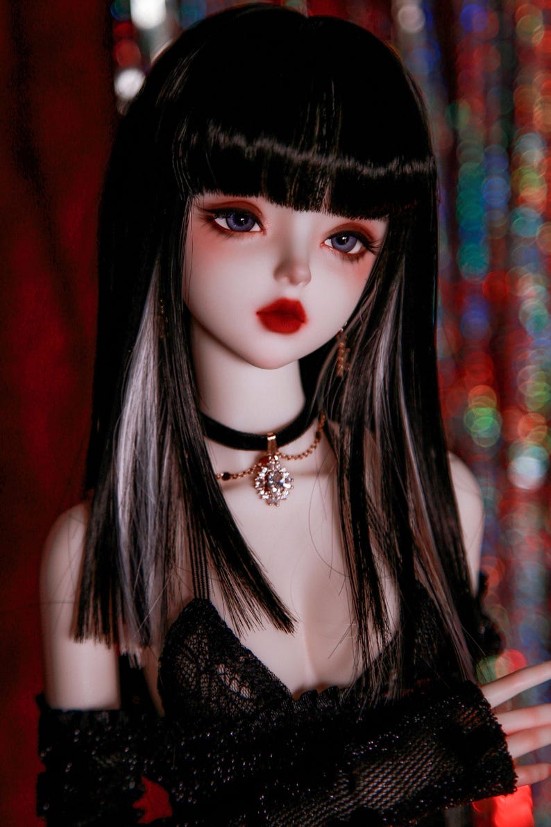 May include: A doll with long black hair with white streaks, wearing a black lace top and a black choker with a rhinestone pendant.