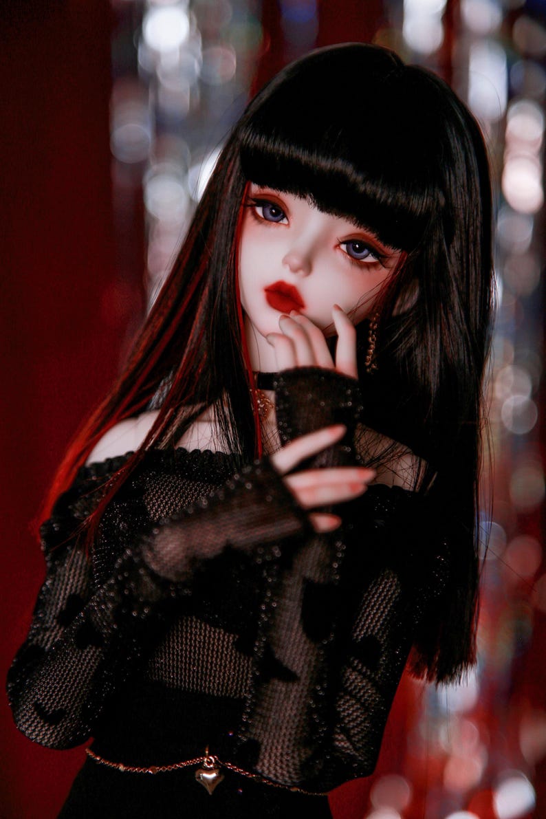 May include: A doll with long black hair with red streaks, wearing a black mesh top with a heart-shaped pendant on a gold chain. The doll has a red lip and blue eyes.