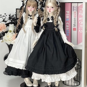 May include: Two dolls wearing black and white dresses with ruffled hems. The dolls have long blonde braids and are wearing black shoes. The dolls are standing on a white surface in front of a bookshelf.