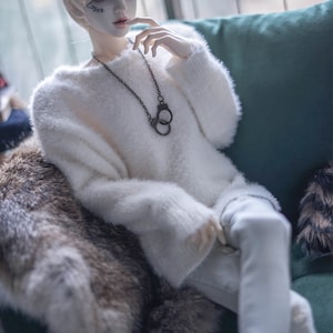 May include: A doll with pale skin, short white hair, and a white fluffy sweater. The doll wears a black handcuff pendant necklace and has the word "Loser" tattooed on its face. The doll is seated on a green sofa with a fur throw.