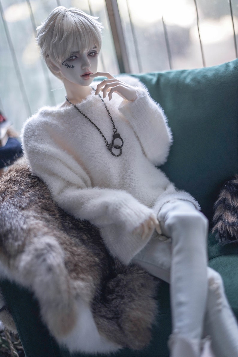 May include: A doll with pale skin, short white hair, and a white fluffy sweater. The doll wears a black handcuff pendant necklace and has the word "Loser" tattooed on its face. The doll is seated on a green sofa with a fur throw.