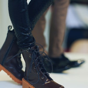 May include: Close-up of a pair of brown leather boots with a lace-up design and a tan sole. The boots have a block heel and are paired with black textured leggings. The background is blurred, showing another person's legs and shoes.