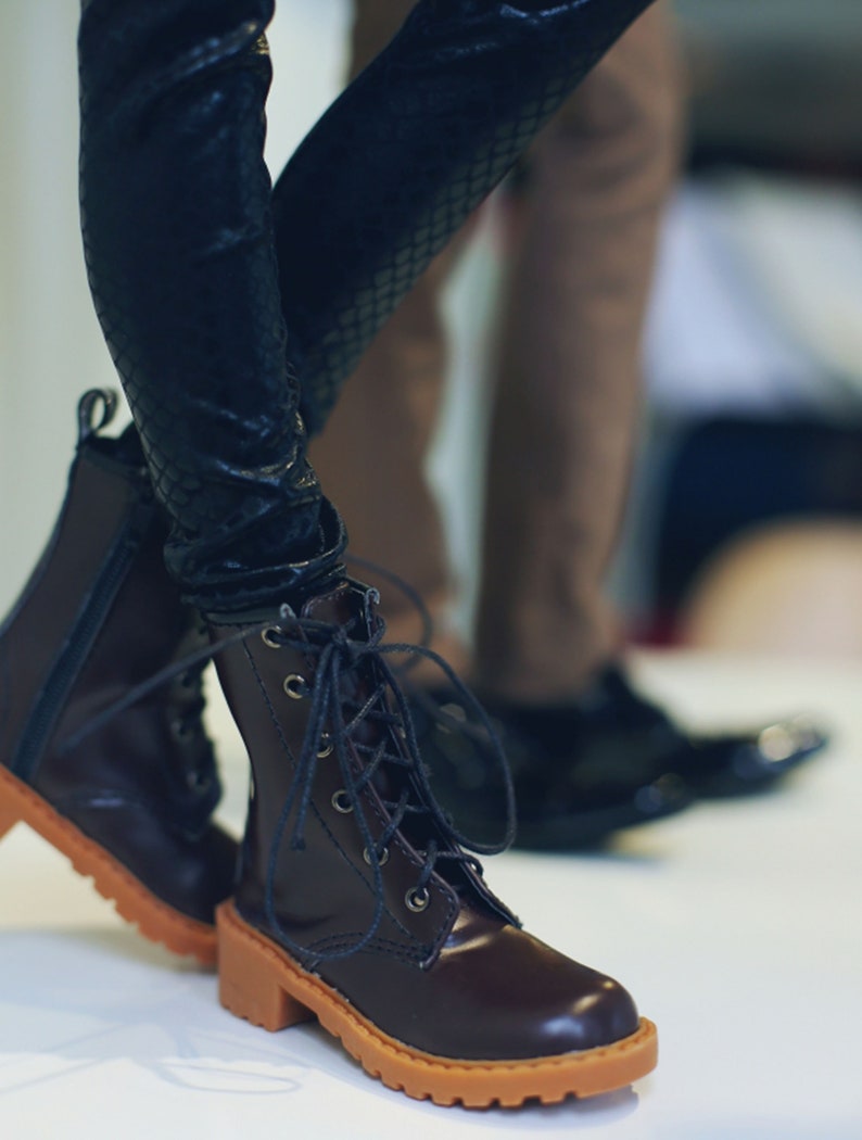 May include: Close-up of a pair of brown leather boots with a lace-up design and a tan sole. The boots have a block heel and are paired with black textured leggings. The background is blurred, showing another person's legs and shoes.