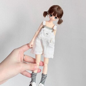 May include: A doll with brown hair in pigtails, wearing a white overall shorts set with the text "TWW" on the bib. The doll has blue eyes and is wearing black and white striped socks and white sneakers with black stripes.