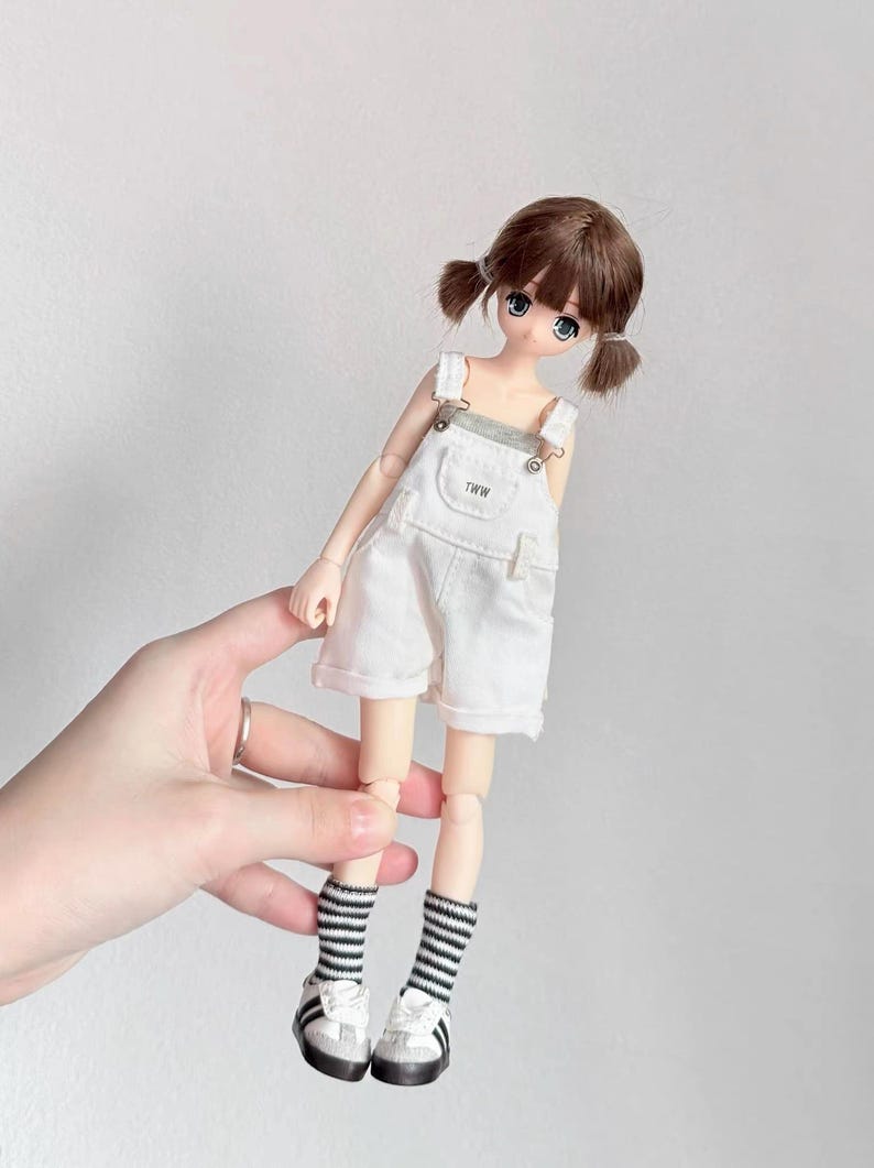 May include: A doll with brown hair in pigtails, wearing a white overall shorts set with the text "TWW" on the bib. The doll has blue eyes and is wearing black and white striped socks and white sneakers with black stripes.