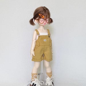 May include: A doll wearing a mustard yellow overall shorts set with a white top. The doll has brown hair in pigtails, round glasses, and striped socks. The doll is wearing white sneakers with black stripes.