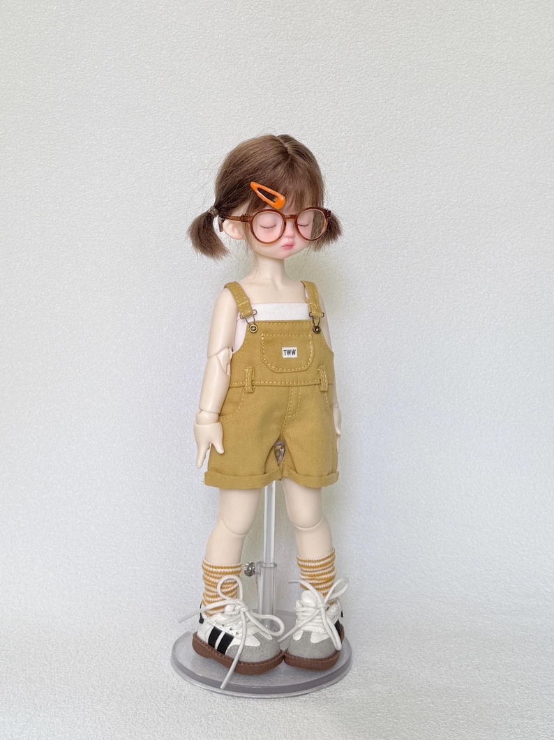 May include: A doll wearing a mustard yellow overall shorts set with a white top. The doll has brown hair in pigtails, round glasses, and striped socks. The doll is wearing white sneakers with black stripes.