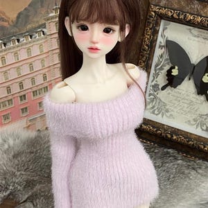 Puede incluir: Una mu&ntilde;eca con un su&eacute;ter rosa claro y peludo de hombros descubiertos. La mu&ntilde;eca tiene el pelo largo y casta&ntilde;o y lleva una falda blanca.