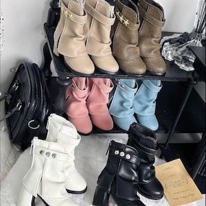 Könnte beinhalten: Eine Kollektion von Stiefeletten in verschiedenen Farben wie Beige, Braun, Rosa, Hellblau, Weiß und Schwarz. Jedes Paar verfügt über einen seitlichen Reißverschluss, einen dekorativen Riemen mit metallischen Akzenten und einen Blockabsatz.
