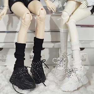 May include: Black and white sneakers with thick soles and patterned laces. The sneakers are paired with black socks. A second pair of white sneakers with patterned laces and white socks are also visible.