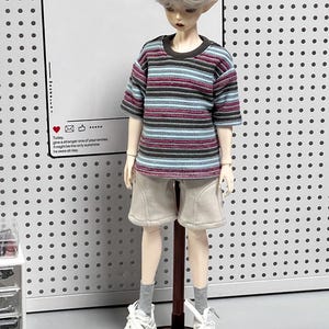 Pu&ograve; includere: Una bambola che indossa una maglietta a righe blu, marrone e rosa, pantaloncini beige, scarpe da ginnastica bianche e calzini bianchi. La bambola &egrave; in piedi su un supporto nero con un palo marrone. La bambola ha i capelli biondi e gli occhi azzurri. Lo sfondo &egrave; un pannello forato bianco.