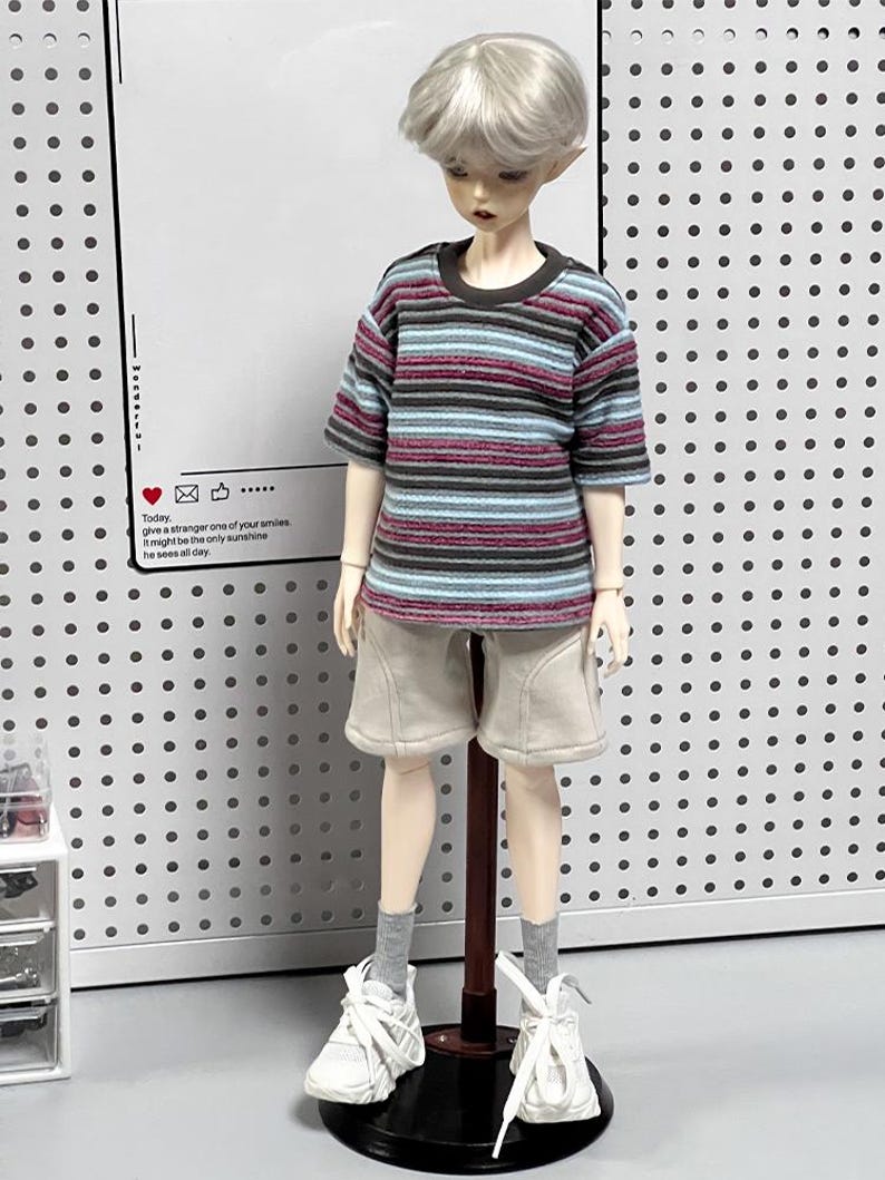 Pu&ograve; includere: Una bambola che indossa una maglietta a righe blu, marrone e rosa, pantaloncini beige, scarpe da ginnastica bianche e calzini bianchi. La bambola &egrave; in piedi su un supporto nero con un palo marrone. La bambola ha i capelli biondi e gli occhi azzurri. Lo sfondo &egrave; un pannello forato bianco.
