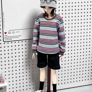 Pu&ograve; includere: Una bambola che indossa una camicia a maniche lunghe a righe, pantaloncini neri, scarpe da ginnastica bianche e un berretto di lana grigio con occhiali rotondi. La bambola &egrave; in piedi su un supporto di legno su uno sfondo bianco con pois neri.