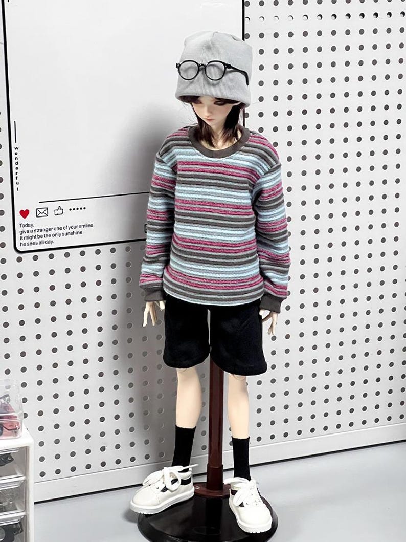 Pu&ograve; includere: Una bambola che indossa una camicia a maniche lunghe a righe, pantaloncini neri, scarpe da ginnastica bianche e un berretto di lana grigio con occhiali rotondi. La bambola &egrave; in piedi su un supporto di legno su uno sfondo bianco con pois neri.