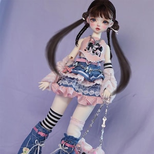 May include: A doll with long black hair styled in pigtails, wearing a pink and blue outfit with a denim skirt, a pink top with a cartoon dog design, and a silver chain necklace. The doll is wearing white socks with black stripes and blue denim boots.