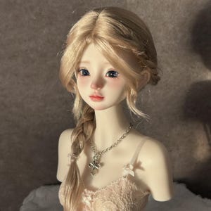Op de afbeelding: Een kogelgewrichtpop met een bleke huid, grote donkere ogen en lichtblond haar in een zijvlecht. De pop draagt een zilverkleurige ketting met een kruishanger en een lichtgekleurde kanten top met dunne schouderbandjes en kleine strikjes.