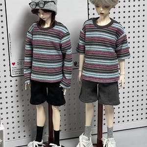 Pu&ograve; includere: Due bambole che indossano camicie a righe e pantaloncini. La bambola a sinistra indossa un berretto grigio e occhiali. Entrambe le bambole sono in piedi su supporti in legno.