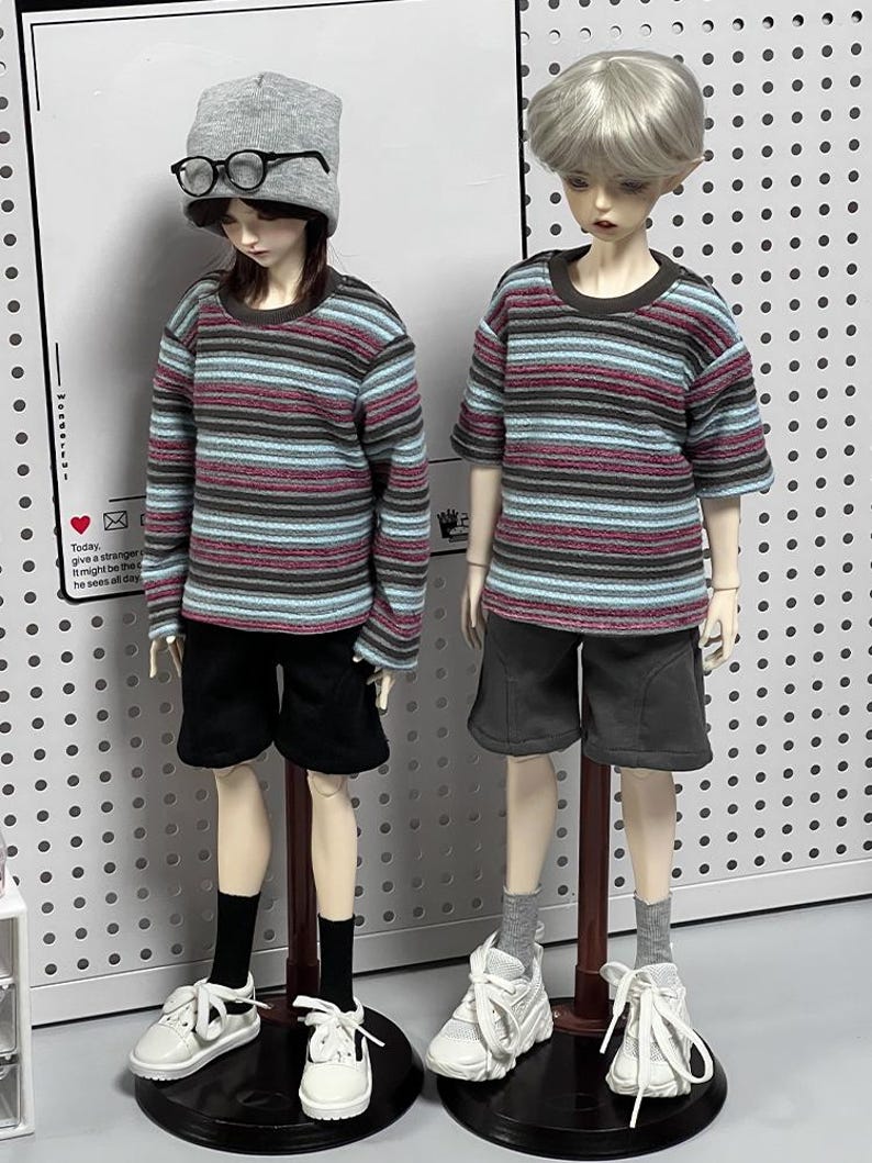 Pu&ograve; includere: Due bambole che indossano camicie a righe e pantaloncini. La bambola a sinistra indossa un berretto grigio e occhiali. Entrambe le bambole sono in piedi su supporti in legno.