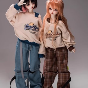 May include: Two dolls dressed in similar beige sweatshirts with "Sun Light" text and a flower design. One doll is wearing blue jeans, the other brown plaid trousers. Both dolls have long hair and stand against a grey backdrop.