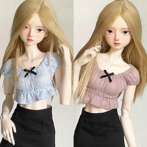 Puede incluir: Dos muñecas con blusas de colores diferentes y faldas negras. La muñeca de la izquierda lleva una blusa azul claro con un lazo negro. La muñeca de la derecha lleva una blusa rosa con un lazo negro.