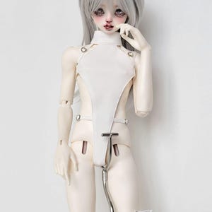 May include: A white doll with silver hair wearing a white, sleeveless, one-piece outfit with straps and metal accents. The doll is standing on a metal stand with a white strap around its leg.
