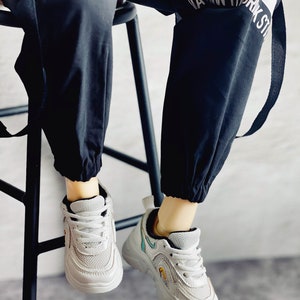 Puede incluir: Zapatillas blancas con detalles negros y dise&ntilde;o de malla. Las zapatillas tienen una suela gruesa y las lleva una persona que viste pantalones negros con una raya blanca y el texto "ATM THRK ST" en el lateral.