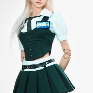 May include: A doll wearing a dark green school uniform with a pleated skirt, a white collared shirt, and a black tie. The doll has a black cap with a silver chain and a name tag. The text "EVERY ORDINARY FACE HIDES AN INTERESTING SOUL" is at the top.