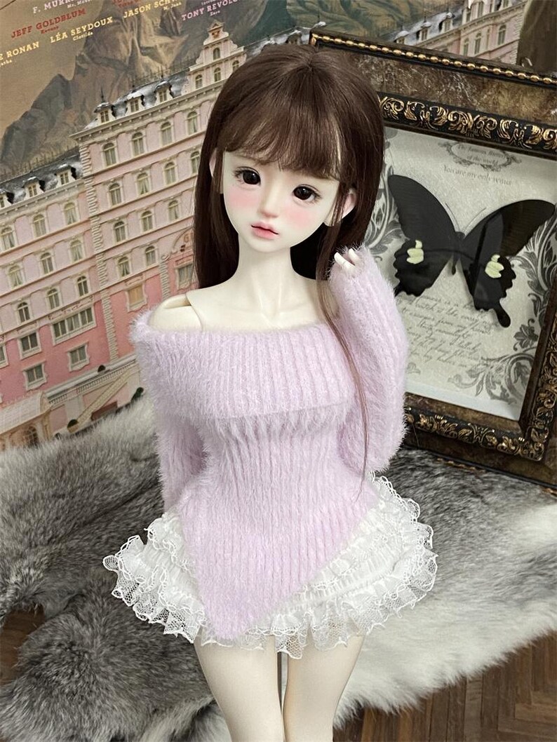 Puede incluir: Una mu&ntilde;eca con un su&eacute;ter de color morado claro y peludo con un borde de encaje blanco. La mu&ntilde;eca tiene el pelo largo y casta&ntilde;o y lleva una falda blanca.