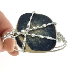May include: A silver bracelet with a black and tan stone set in the center. The stone is surrounded by a silver wire design that resembles a branch.