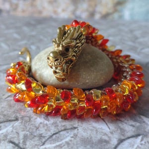 May include: A bracelet featuring a metallic gold dragon head focal piece with an open mouth, connected to a flexible band constructed from small, translucent red, orange, and yellow glass beads arranged in a dense, overlapping scale-like pattern.