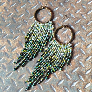 Puede incluir: Un par de pendientes hechos a mano con aros color latón y flecos de cuentas. Los flecos presentan una mezcla de cuentas verdes, amarillas, blancas y negras. Los pendientes están diseñados para orejas perforadas.
