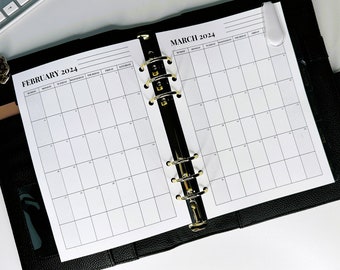 Puede incluir: Una agenda encuadernada en cuero negro con anillos dorados. La agenda tiene dos páginas abiertas con calendarios mensuales para febrero y marzo de 2024.
