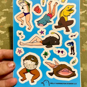 May include: A sticker sheet with various cartoon sea creatures in swimsuits and sunglasses on a blue background. The sticker sheet includes a fish, shark, and other sea creatures. The text "MOLLY HARRINGTON STUDIOS LLC" is at the bottom.