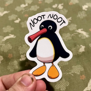 Puede incluir: Una pegatina con un pingüino de dibujos animados con las palabras "NOOT NOOT" encima. El pingüino tiene un cuerpo negro, vientre blanco, pies naranjas y un pico rojo. La pegatina está sobre un fondo blanco.